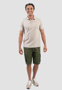 Polo beige con maniche corte e rifiniture azzurre, abbinato a pantaloni cargo verdi e scarpe da ginnastica chiare. In piedi contro uno sfondo neutro.