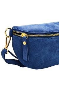 Sac en suede bleu avec une fermeture éclair dorée, de forme arrondie, bandoulière et une petite étiquette en tissu pour la tirette. Gros plan sur la fermeture éclair et les détails du matériel.