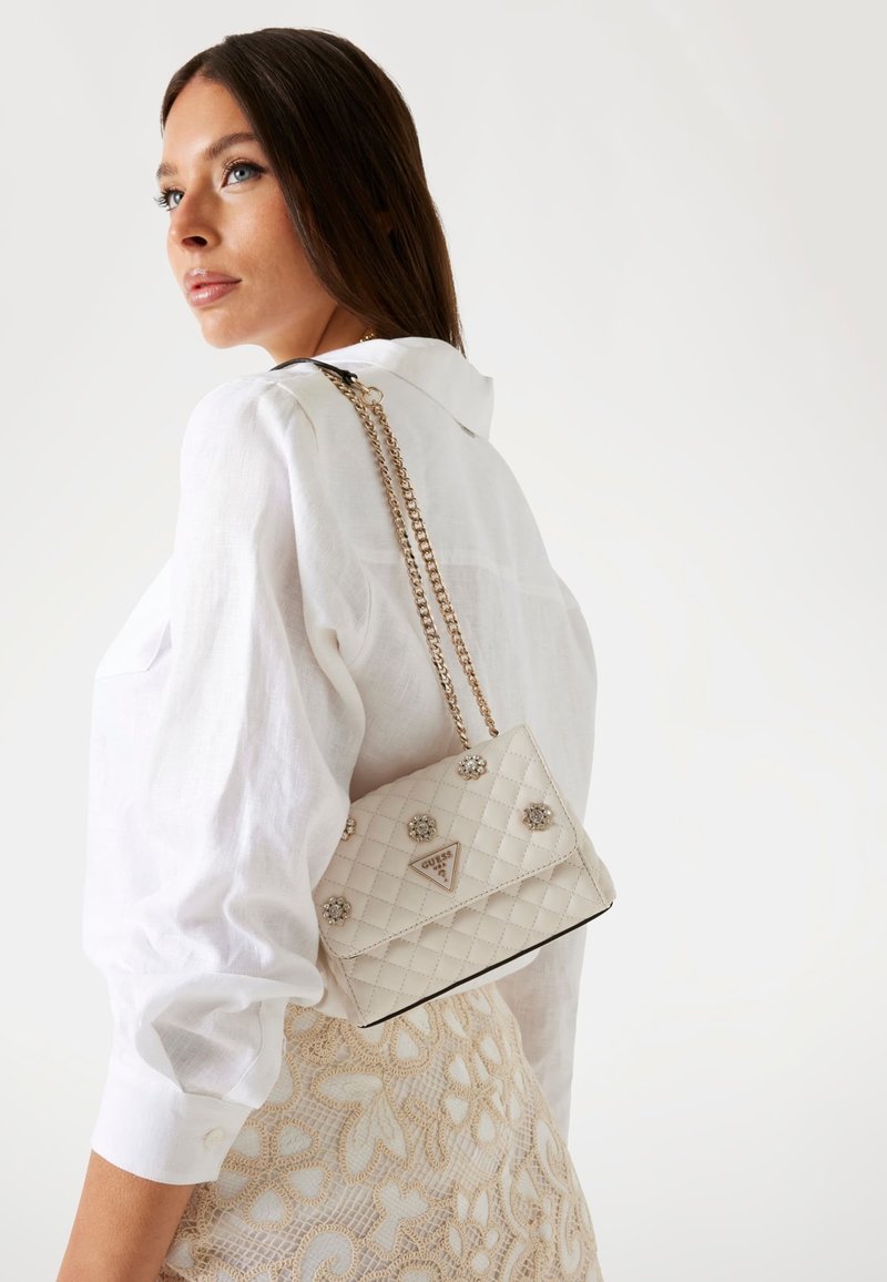 Borsa beige trapuntata con tracolla a catena, decorata con ornamenti in argento, esposta su uno sfondo bianco.