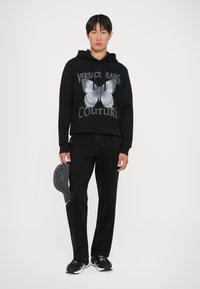 Hanorac negru cu grafică de fluture și text "VERSACE JEANS COUTURE", purtat cu pantaloni negri, ținând o șapcă gri, cu pantofi sport negri.