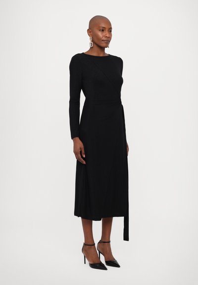 Vestido negro ajustado con mangas largas, de textura suave, hasta la rodilla, con un detalle de lazo lateral. Combinado con tacones negros de punta afilada.