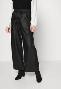 Pantalones anchos y negros hechos de un material de piel sintética suave. Cuentan con una cinturilla elástica y un ajuste relajado. Sin patrones ni acentos visibles.