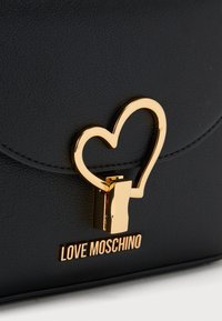 Μαύρη τσάντα χειρός από ανάγλυφο δέρμα με χρυσό κλιπ σε σχήμα καρδιάς και χρυστό λογότυπο "LOVE MOSCHINO" στο κάτω μέρος.