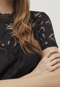 Top in pizzo nero con motivi floreali, scollatura a scallop e maniche corte. La texture è semi-trasparente con un aspetto tattile.