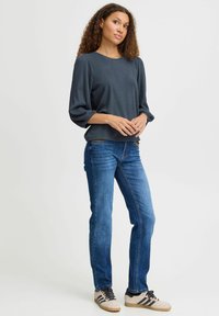 Maglietta a maniche lunghe a costine grigia scura abbinata a jeans in denim blu. La maglietta ha una vestibilità comoda e maniche voluminose; le scarpe sono color crema con strisce nere.