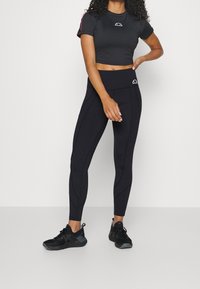 Top corto negro con mangas cortas, que presenta un pequeño logo y acentos rosas en los laterales; combinado con leggings negros con un diseño de costura texturizada.