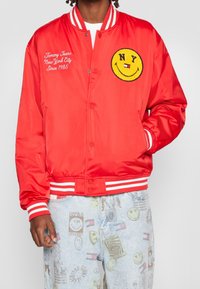 Giacca bomber in satin rosso con polsini e vita a strisce bianche. Presenta un faccina sorridente gialla ricamata e del testo sul lato sinistro del petto.