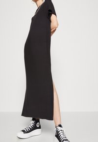 Czarna maxi sukienka z krótkimi rękawami i bocznym rozcięciem, wykonana z miękkiego materiału, w połączeniu z czarnymi butami sportowymi typu high-top. Prosta, prosta sylwetka.