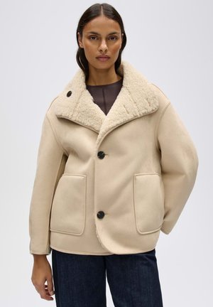 Frau trägt eine beige Schaffelljacke mit großem Kragen und zwei Fronttaschen, kombiniert mit dunklen Jeans, steht vor einem einfarbigen Hintergrund.