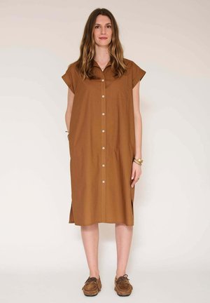 Femme debout portant une robe chemise marron ample à manches courtes et des mocassins marron, sur un fond clair uni.