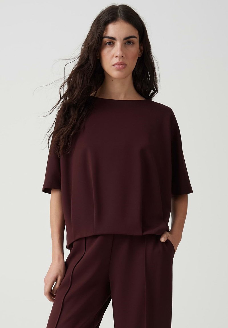 Woman with long dark hair wears loose dark burgundy short-sleeve top and matching pants, standing with one hand in pocket against plain background.