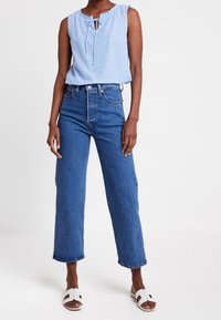 Kvinna i en ärmlös blå randig topp, hög midja med vida blå jeans och vita sandaler som står på en vit bakgrund.