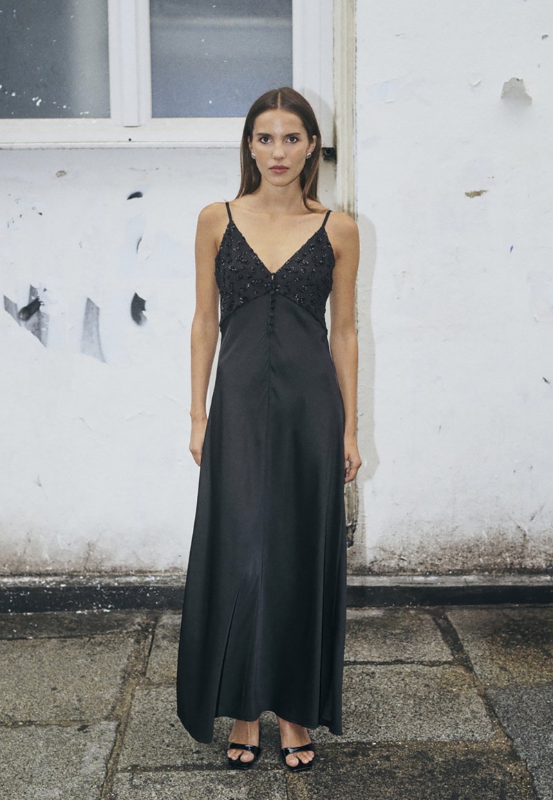 Robe maxi en satin noir avec un décolleté profond en V et des embellissements perlés. Elle présente des bretelles fines et une fente latérale, complétée par des sandales noires à lanières.