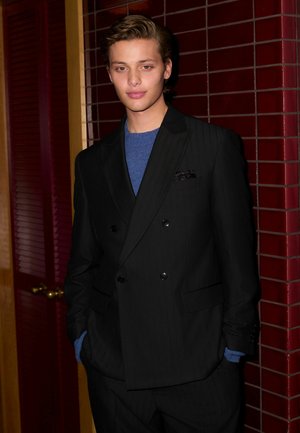 Schwarzer doppelt geknöpfter Anzug mit vertikalen Nadelstreifen, kombiniert mit einem blauen Pullover und einer Tasche mit Einstecktuch. Vor einer wand mit kastanienbraunen Fliesen.