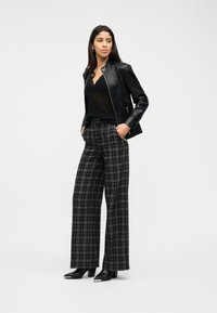 Γυναίκα που φορά μαύρο δερμάτινο μπουφάν πάνω από μαύρο τοπ, καρό παντελόνι φαρδιάς γραμμής και μαύρες μποτάκια με τακούνι, στέκεται με τα χέρια στις τσέπες.