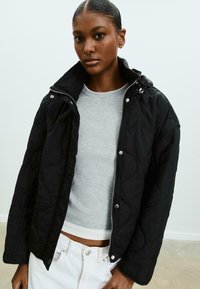 Veste noire matelassée avec un col montant, fermeture éclair argentée et cordon de serrage. Portée sur un haut en maille gris clair et un jean blanc.