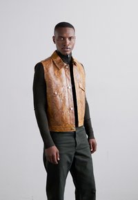 Gilet en cuir marron avec poches avant, boutons en argent et texture lisse, porté par-dessus un haut noir à manches longues et un pantalon vert foncé.