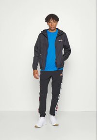 Schwarze leichte Jacke, blaues T-Shirt, schwarze Hose mit buntem Seitenmuster und weiße Sneaker. Das Modell steht vor einem einfarbigen Hintergrund.