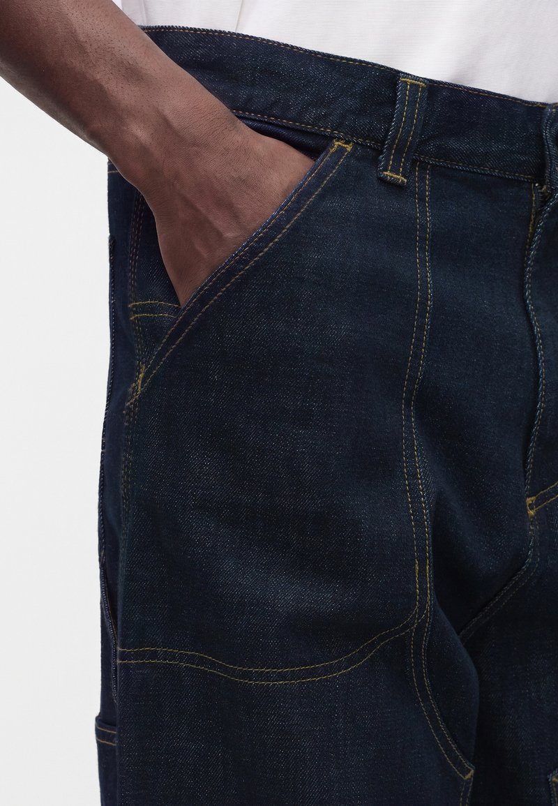 Hand resting inside the front pocket of dark blue denim jeans with visible yellow stitching and a white shirt tucked in.