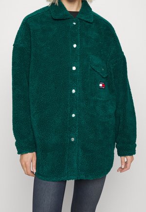 Personne portant une veste oversize en polaire sherpa verte avec des boutons-pression argentés et une poche poitrine avec un logo Tommy Jeans.