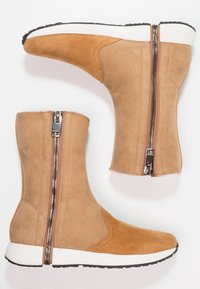 Botas de tobillo de ante marrón claro con una suela blanca, que cuentan con un cierre lateral y acentos marrones en contraste. Textura suave, diseño minimalista.