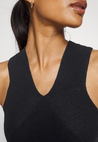 Débardeur noir en maille côtelée avec un col en V, présentant des motifs géométriques et des détails texturés aux épaules.