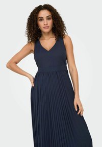 Navy pleated maxi dress with a V-neckline, sleeveless design, textured fabric, and flared bottom. No visible hardware or accents.