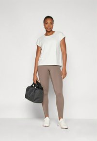 Ljusgrå kortärmad topp tillsammans med bruna leggings; modell håller en svart sportväska; vita atletiska skor med lila detaljer.