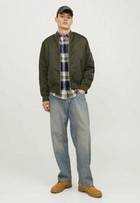 Chaqueta bomber en verde oliva, camisa de cuadros con botones, jeans ligeros y sueltos en azul claro y calzado marrón. La chaqueta tiene bolsillos con cremallera.
