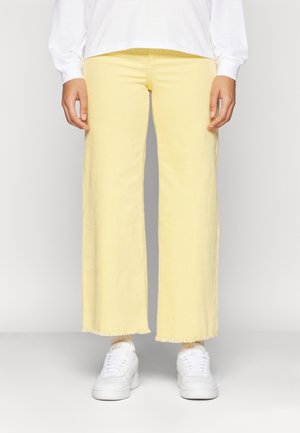 Person wearing light yellow wide-leg corduroy pants, white long-sleeve top, white socks, and white sneakers, standing against a plain background.