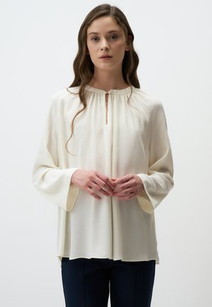 Blusa crema con scollatura tonda e dettaglio a occhio di serratura, caratterizzata da maniche lunghe e ampie e una texture morbida e fluida. Abbinata a pantaloni scuri.