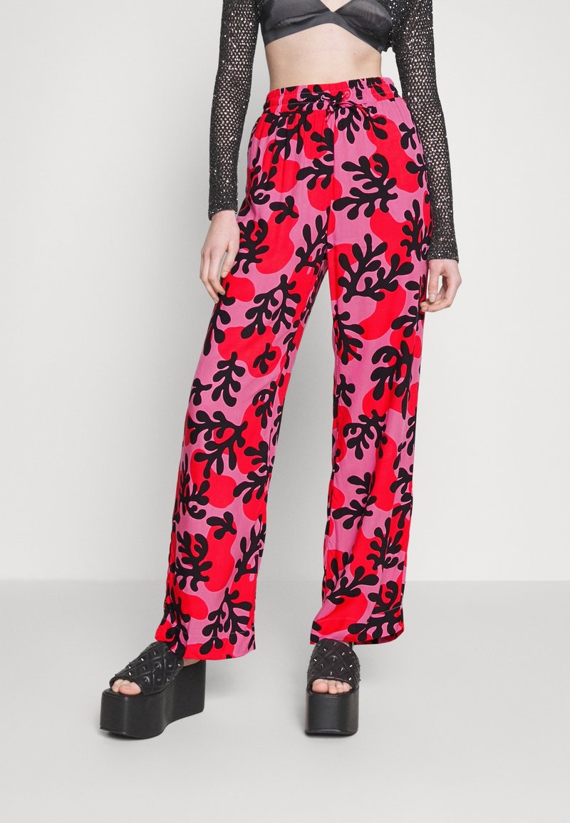 Femme portant un pantalon taille haute à jambes larges avec un motif abstrait de feuilles rouges, roses et noires, ainsi que des sandales compensées noires.