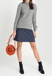 Grauer gestrickter Pullover mit strukturiertem Muster, rundem Ausschnitt und dekorativen Bindebändern an den Manschetten, kombiniert mit einem marineblauen Wildlederrock und einer orangen runden Handtasche.