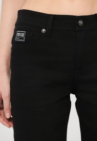 Black denim jeans with a slim fit, silver hardware, and a visible brand label on the waistband. Features contrast stitching and five pockets.