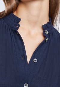Granatowa bluzka z teksturowanej tkaniny, z falbaniastym dekoltem, plisą z trzema guzikami oraz subtelnym wzorem w jodełkę.