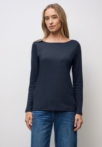 Marineblaues Langarmoberteil mit einem Bootsausschnitt, gefertigt aus weichem, glattem Stoff. Kombiniert mit hellblauen Jeans, minimaler Struktur und Detail.