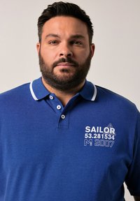 Mörkblå polo med vita accenter och tre knappar, med tryckt text: "SAILOR 53.281534 M: 2007" på vänster bröst.