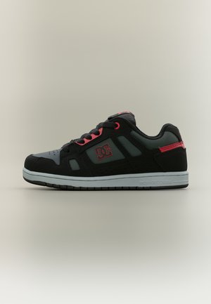 Juodos ir pilkos spalvos žemo profilio sportbačiai su raudonomis detalėmis, juodais raišteliais ir DC logotipu šone, pateikti ant neutralios, vientisos spalvos fono.
