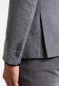 Szary wełniany blezer o dopasowanym kroju, widocznych przeszyciach oraz sześciu ciemnych guzikach na mankiecie. Gładka faktura i wyraźnie zarysowany krój.