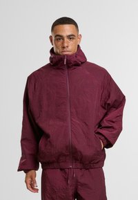 Bordowa kurtka z kapturem, zapinana na zamek, o teksturowanej tkaninie, z regulowanym dołem i bocznymi kieszeniami, zaprojektowana z pikowanymi wzorami.