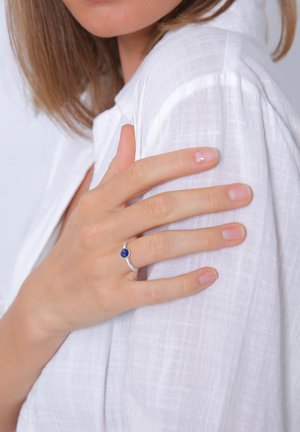 Silberner Ring mit einem runden blauen Stein, getragen an einer Hand, die auf einem weißen, strukturierten Stoff ruht. Saubere Linien und minimalistisches Design heben den Stein hervor.