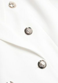 Witte stof met een gladde textuur, voorzien van opvallende metalen knopen met reliëfontwerpen, die een dubbelbreasted stijl laten zien.