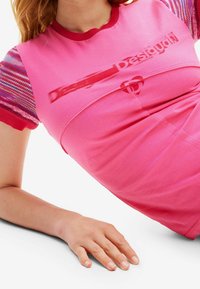 Růžové tričko s krátkými rukávy v různých barvách s pruhy, žebrovaným výstřihem a výrazným logem potištěným na přední straně. Jemná textura.