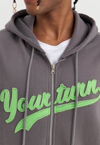 Šedá mikina na zip s kapucí, zdobená zeleným vyšitým nápisem "Tvůj tah" a detaily, vybavená pohodlnou kapucí a strukturovanou látkou.