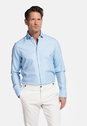 Chemise à manches longues bleu clair avec un col standard, fermeture à boutons et tissu texturé. Associée à un pantalon blanc avec des boutons dorés.