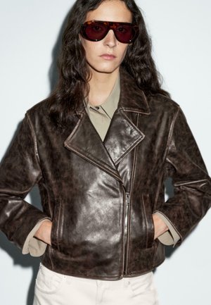 Braune Kunstleder-Bikerjacke mit Reverskragen, Reißverschluss und zwei seitlichen Taschen. Kombiniert mit einem hellgrünen Hemd darunter.