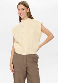 Crème-kleurige gebreide mouwloze trui met textuur kabeldetails, hoge hals en losse pasvorm, gecombineerd met bruine op maat gemaakte broeken.