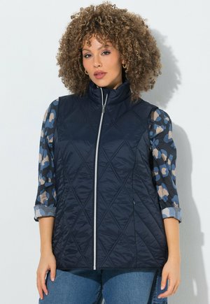 Donna con capelli ricci che indossa un gilet trapuntato blu navy sopra una camicia blu a maniche lunghe con motivi e jeans blu, in piedi davanti a uno sfondo bianco.