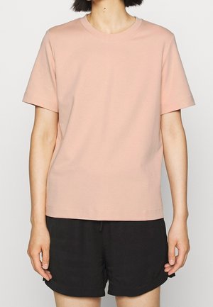 Osoba ubrana w gładki, jasnoróżowy t-shirt z krótkim rękawem i luźne czarne szorty, stojąca na jednolitym tle.