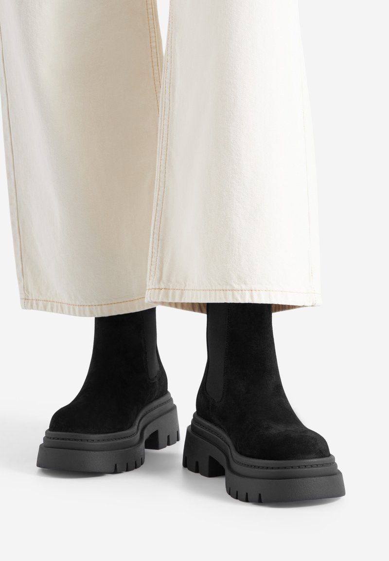 Czarne zamszowe botki za kostkę z grubą gumową podeszwą i elastycznymi panelami po bokach, zestawione z szerokimi kremowymi spodniami, ukazujące teksturowaną powierzchnię.